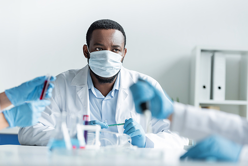 African american scientist in protective mask looking at blurred colleagues working in lab