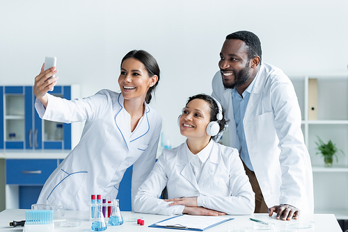 Smiling scientist taking selfie on smartphone near multiethnic colleagues in laboratory