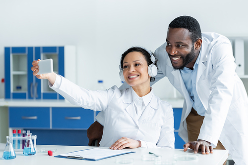 African american scientist in headphones using smartphone near smiling colleague in lab