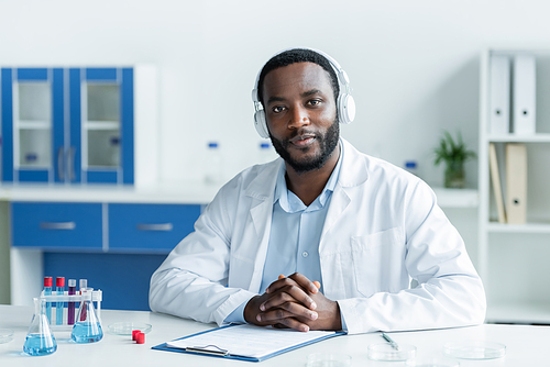 African american scientist in headphones looking at camera near flasks and clipboard