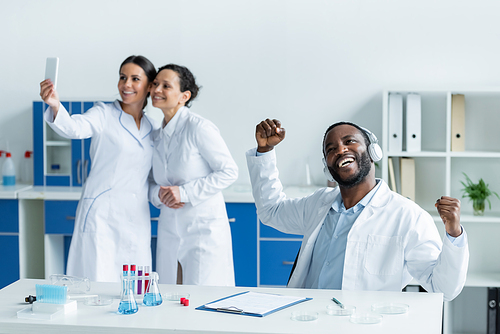 African american scientist in headphones showing yes gesture near blurred colleagues with smartphone in lab