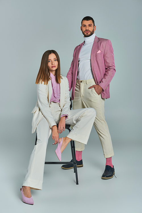 stylish woman in white suit sitting on chair near confident man in lilac blazer on grey, full length