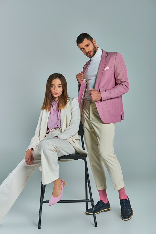 trendy man in lilac blazer looking at camera near young woman in white suit sitting on chair on grey