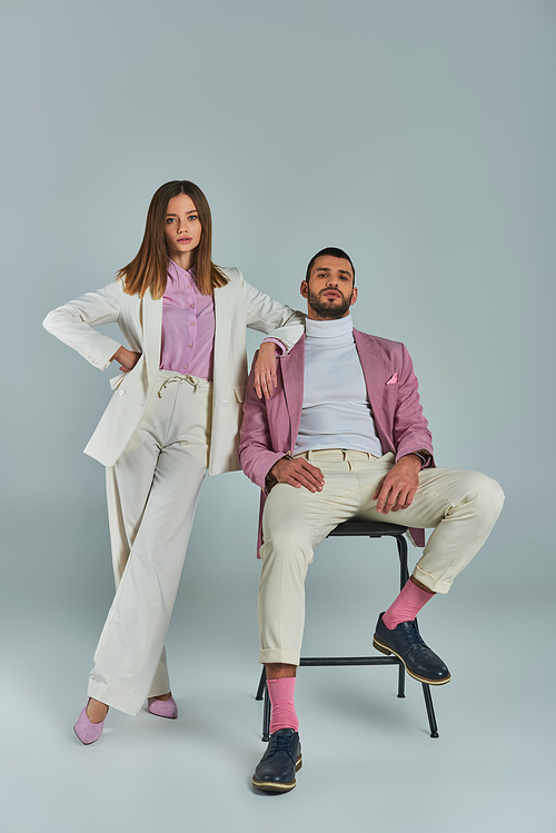 woman in white formal suit posing with hand on hip near man in lilac blazer sitting on chair on grey