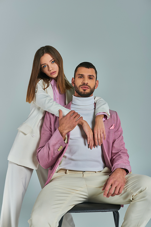trendy woman in white suit embracing confident man in lilac blazer sitting on chair on grey backdrop