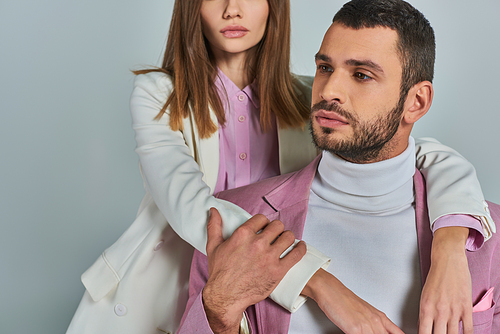 woman in stylish business clothes embracing thoughtful man in lilac blazer looking away on grey