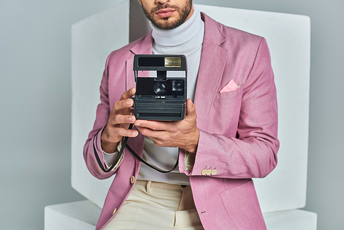 cropped view of modern man in lilac blazer taking photo on vintage camera near white cubes on grey
