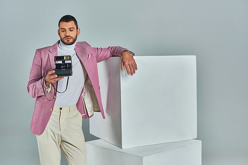 young man in stylish formal wear posing near white cubes with vintage camera on grey, modern fashion