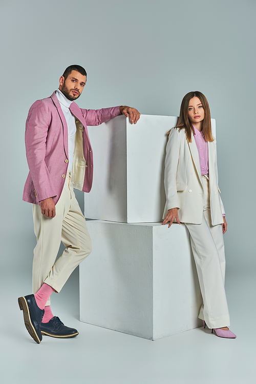 young confident couple in fashionable suits posing near white cubes on grey, minimalistic fashion