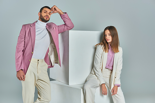 trendy man in lilac blazer looking at camera near white cubes and stylish woman sitting on grey