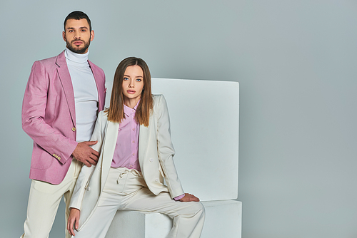 woman in white suit and man in lilac blazer looking at camera near white cubes on grey, trendy wear