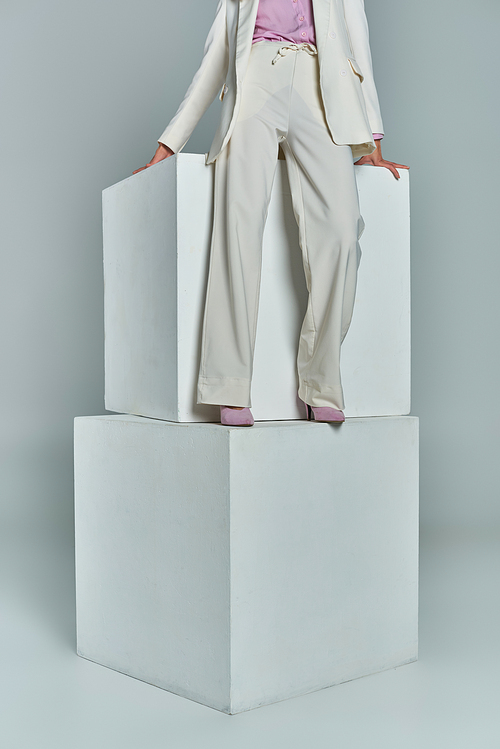 cropped view of young woman in elegant formal attire standing on white cubes on grey backdrop