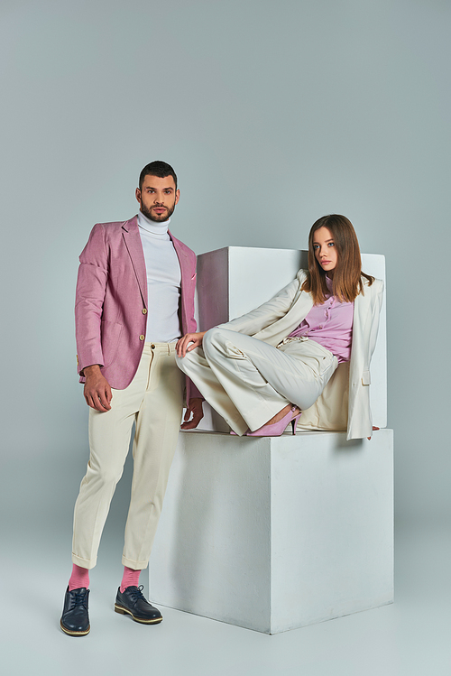 fashionable man in lilac blazer looking at camera near woman in white suit posing on cubes on grey