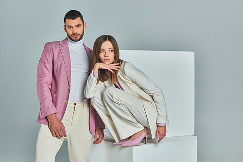 confident man in lilac blazer looking at camera near stylish woman posing on white cubes on grey