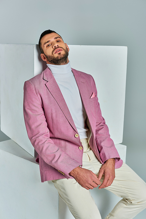 charismatic man in fashionable lilac blazer sitting on white cube and looking at camera on grey