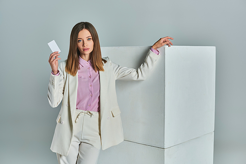 young successful woman in stylish formal attire holding blank business card near white cubes on grey