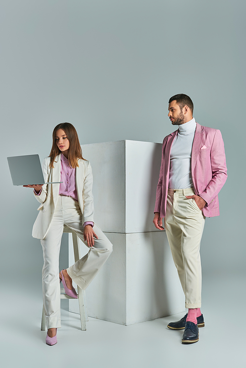 woman in white suit with laptop near man in lilac blazer with hand in pocket next to cubes on grey