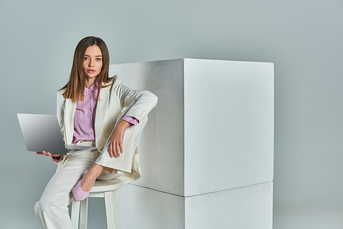 elegant woman in white suit sitting on stool with laptop and looking at camera near cubes on grey