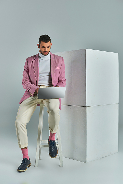 young businessman in lilac blazer sitting on stool and working on laptop near white cubes on grey