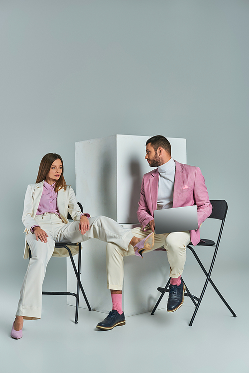 stylish man with laptop and young woman in white suit sitting on chairs near cubes on grey backdrop