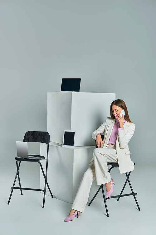 smiling woman sitting on chair and talking on smartphone near devices on white cubes on grey