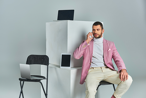 confident stylish man talking on smartphone while sitting near devices on white cubes on grey