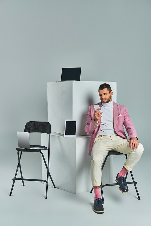 elegant man in lilac blazer sitting on chair with smartphone near devices on white cubes on grey