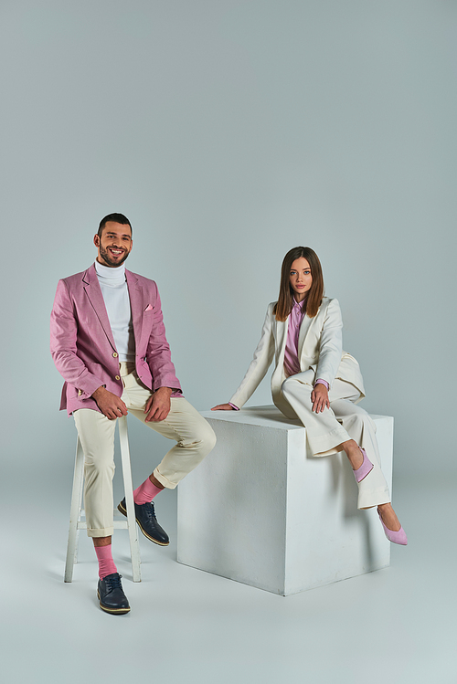 stylish man smiling on chair near woman in suit posing on white cube on grey, business fashion