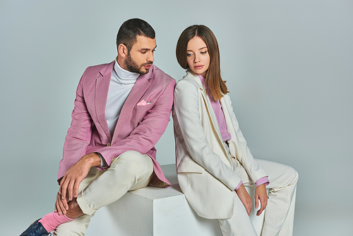 young and fashionable couple in elegant suits sitting on white cube on grey backdrop in studio
