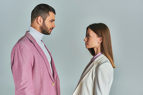 side view of young couple in stylish blazers looking at each other on grey, minimalistic fashion