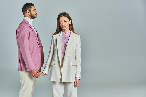 young woman in white suit holding hands with man in lilac blazer and looking at camera on grey