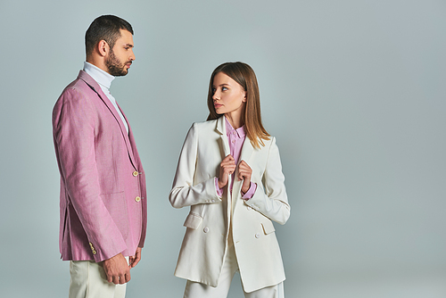 elegant woman in white suit looking at confident man in lilac blazer on grey, modern business couple
