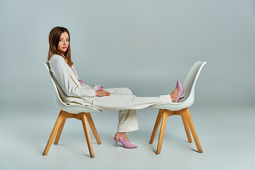 young and elegant woman in white suit posing on armchairs on grey, minimalistic business fashion