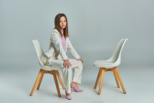 young and stylish woman in white suit sitting on armchair and looking at camera on grey backdrop