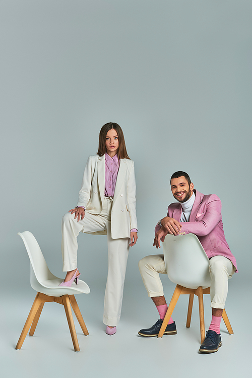 joyful man in lilac blazer sitting and looking at camera near woman posing with armchair on grey
