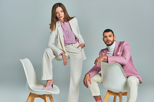 trendy man in lilac blazer sitting near woman in white elegant suit posing with armchair on grey