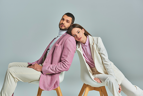 elegant couple in formal wear sitting in armchairs and looking at camera on grey, modern fashion