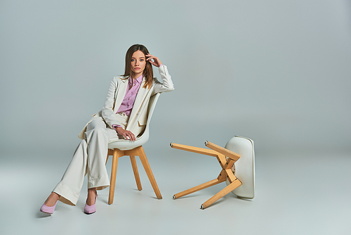 full length of modern woman in white suit sitting on armchair and looking at camera on grey