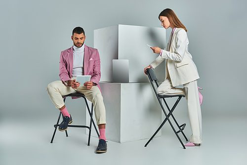 stylish couple using smartphone and digital tablet near laptop on white cubes on grey backdrop