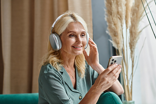 cheerful middle aged woman in wireless headphones listening music and using smartphone at home