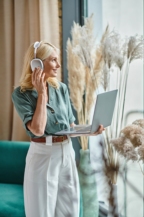 positive middle aged woman in wireless headphones listening music and using laptop at home