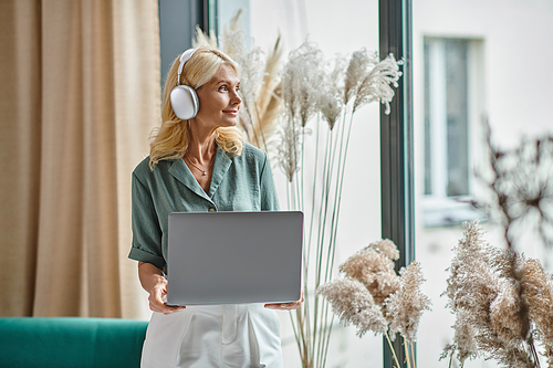 pleased middle aged woman in wireless headphones listening music and holding laptop at home