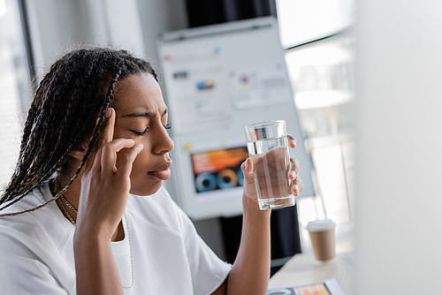 African american businesswoman holding glass of water while suffering from headache in office