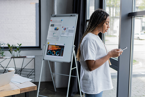 Side view of african american businesswoman using smartphone near window in modern office