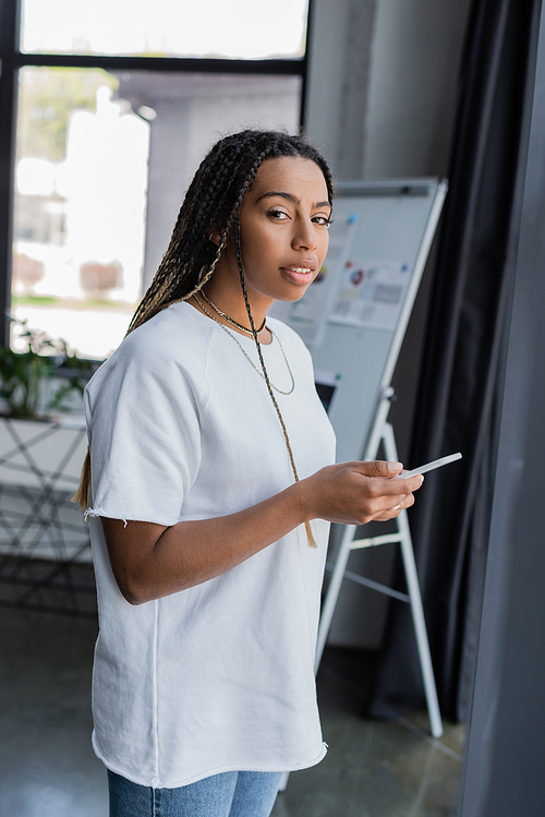 African american businesswoman in casual clothes holding smartphone and looking at camera in office