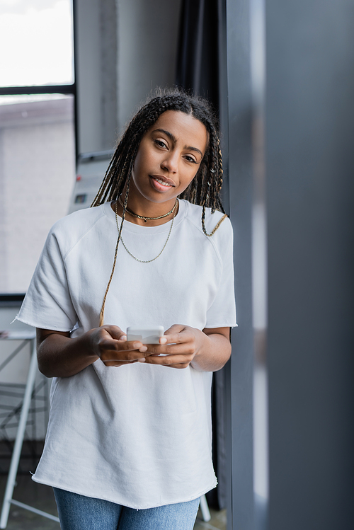 African american businesswoman smiling at camera while holding smartphone in office