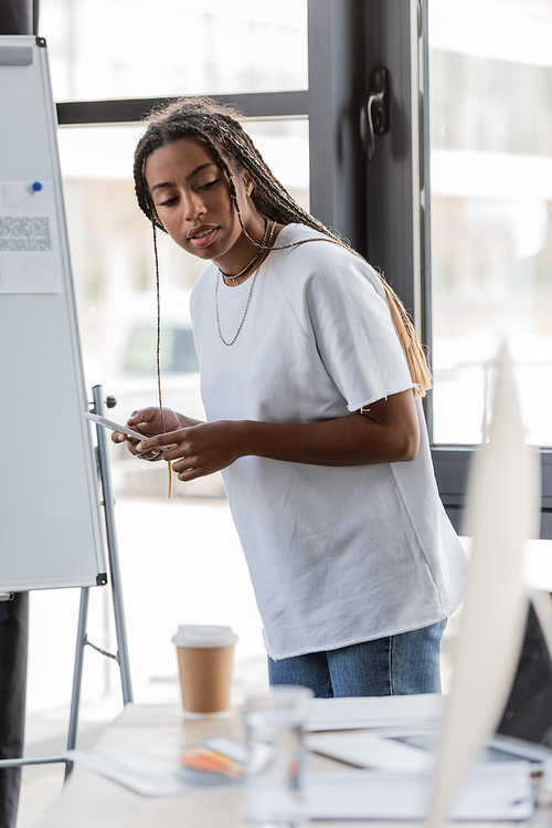 African american businesswoman holding smartphone near blurred laptop and drinks in office