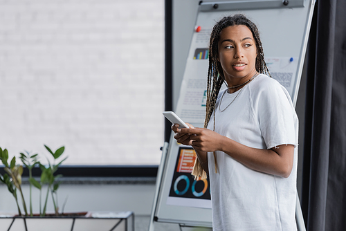 African american businesswoman in t-shirt holding smartphone near blurred flip chart in office