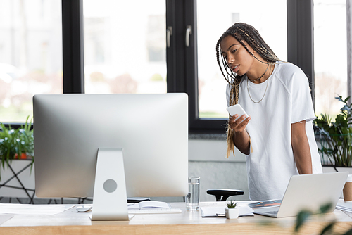 African american businesswoman in t-shirt holding smartphone near computers and papers in office