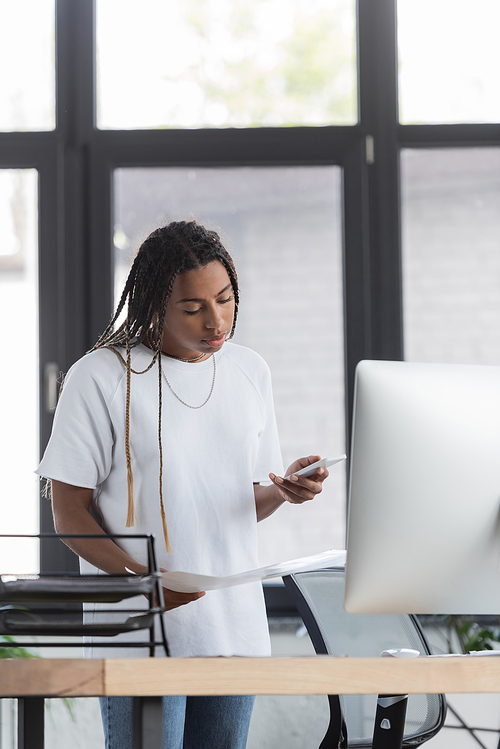 African american businesswoman in casual clothes holding papers and using cellphone in office